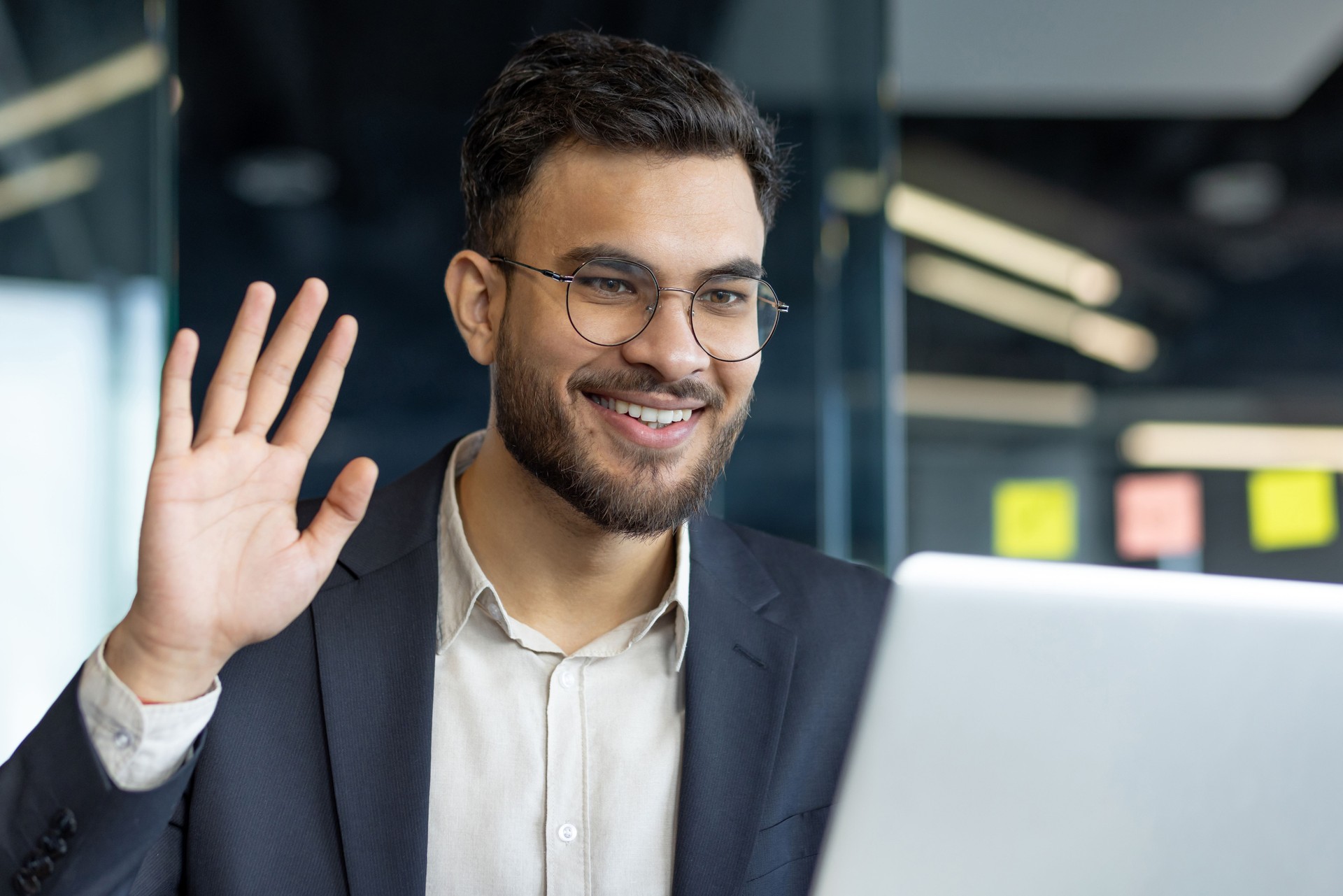 Smiling businessman waving during a video call while working on a laptop in a modern office setting. Confident and friendly interaction in a professional business environment.